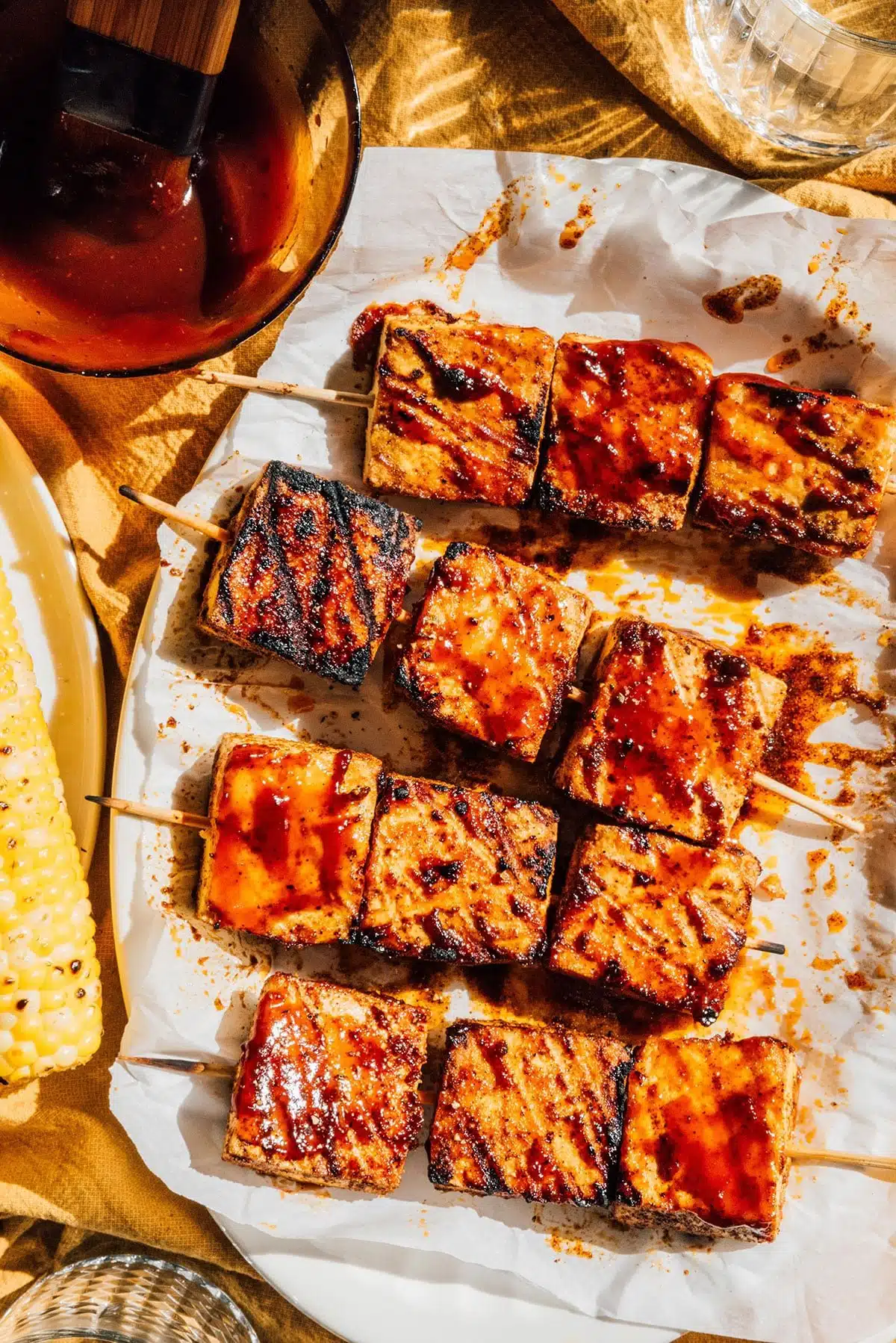 Grilled tofu skewers glazed with a savory sauce, accompanied by charred corn on the cob, all served on a rustic table basked in warm sunlight.