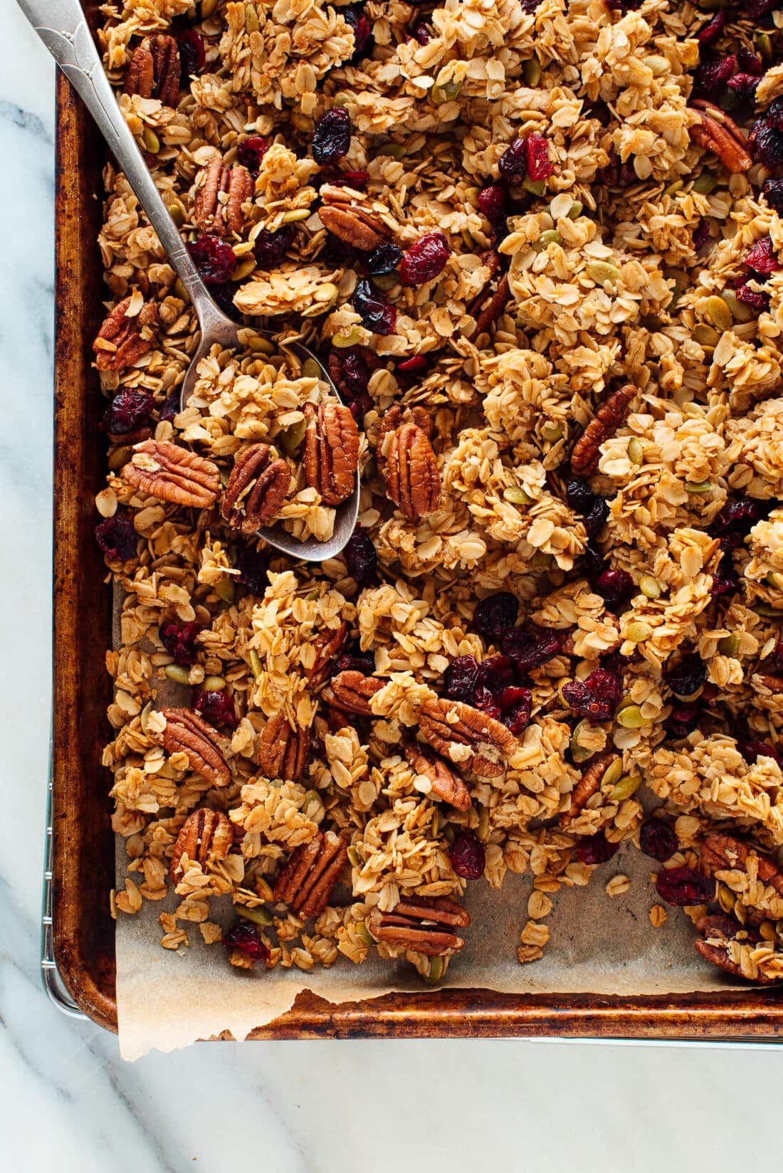 Homemade granola with oats, pecans, and dried cranberries is laid out on a baking sheet.