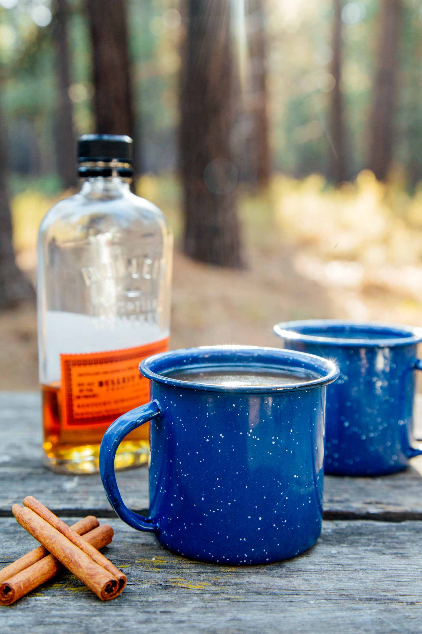 Forest scene with two camping mugs of spiked apple cider with a bottle of bourbon in the background.
