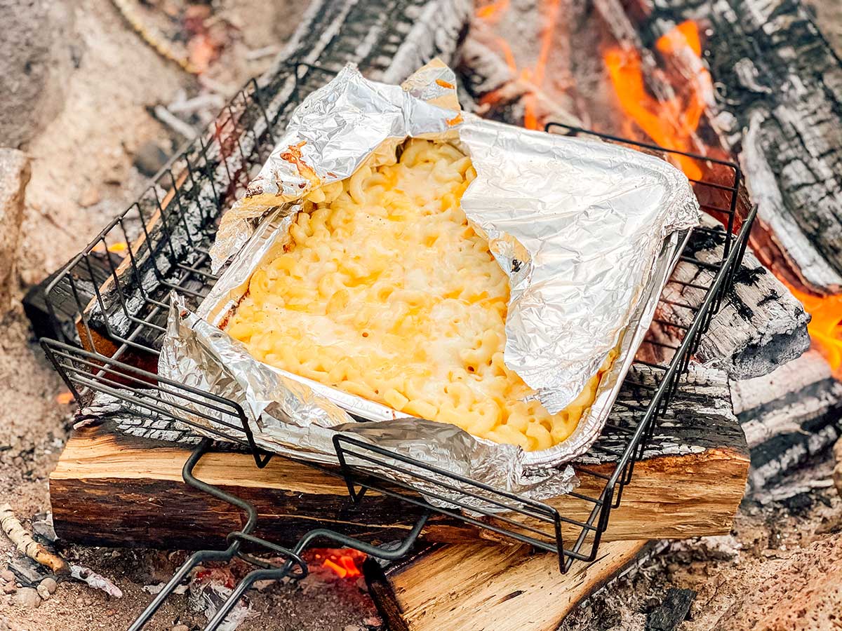 A foil tray of golden macaroni and cheese sits over an open campfire.