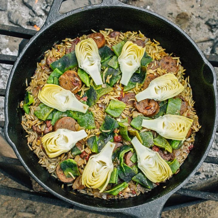 Rice, sausage, artichoke hearts, and poblano pepper arranged in a skillet over a campfire