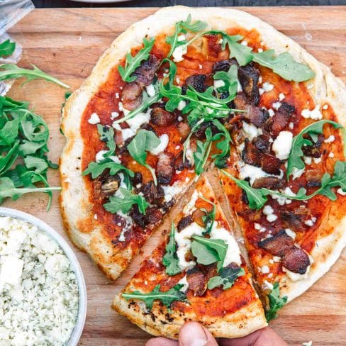 A hand reaching for a slice of freshly made pizza topped with tomato sauce, cheese, arugula, and pieces of bacon on a wooden cutting board.