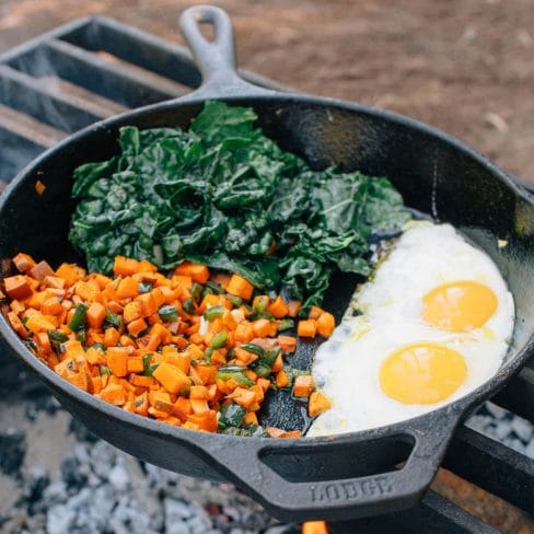 Kale, sweet potato, and two eggs in a skillet over a campfire