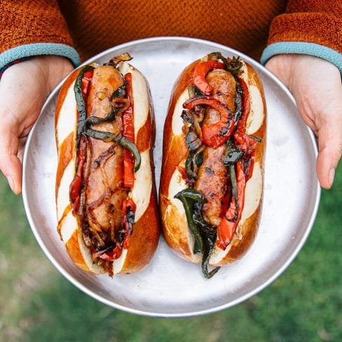 A person holding a plate with two gourmet hot dogs topped with grilled vegetables and sauces.