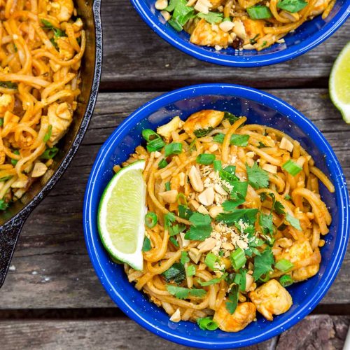 Two bowls of Pad thai and a wedge of lime next to a skillet on a wooden surface