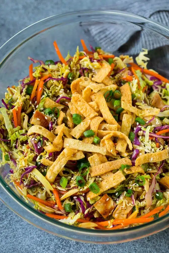 An Asian-inspired salad in a clear glass bowl is garnished with crispy wonton strips, featuring a colorful mix of shredded cabbage, carrots, and sesame seeds.
