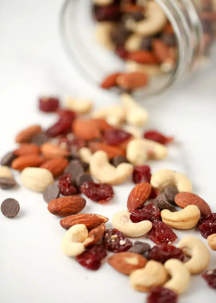 An overturned glass jar has spilled a mix of nuts, dried cranberries, and chocolate chips onto a white surface.
