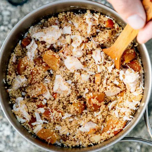 A backpacking pot of couscous, chicken, and dried apricot pieces. A hand is reaching in with a spoon.