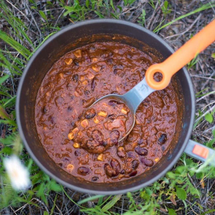 A backpacking pot of chili and a spoon resting in the grass.