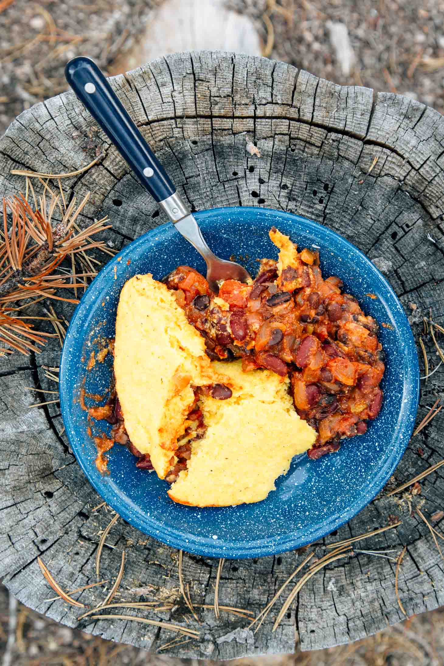Chili and cornbread in a blue camping bowl on a stump.