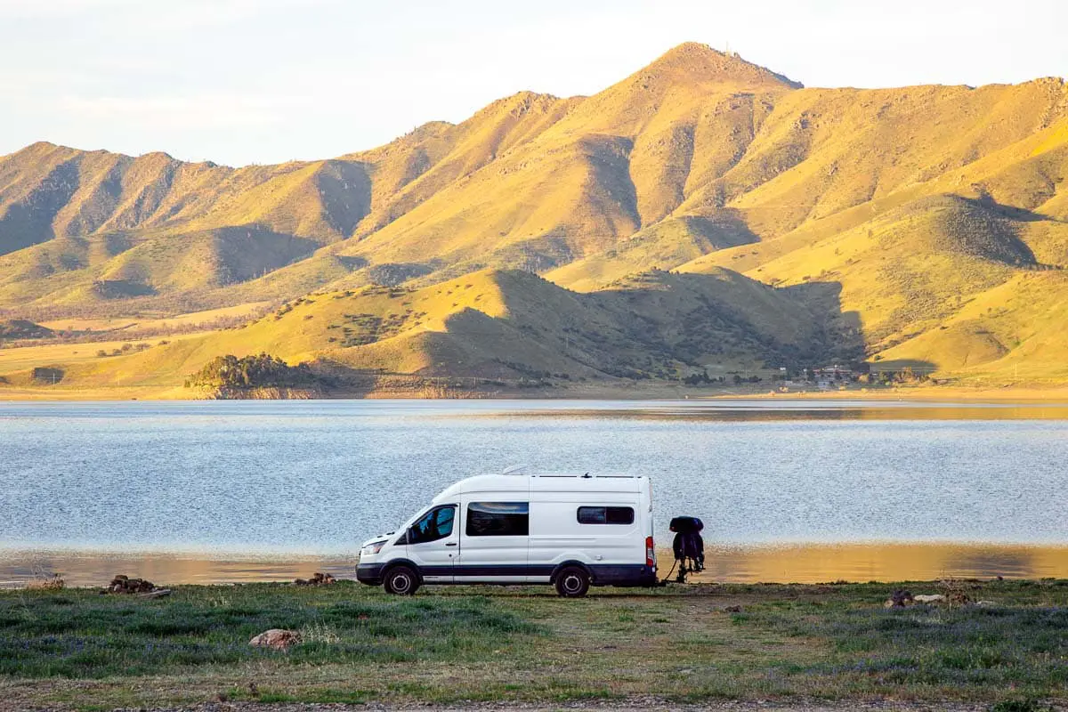 A white camper van parked in front of a lake with a grassy hill in the background