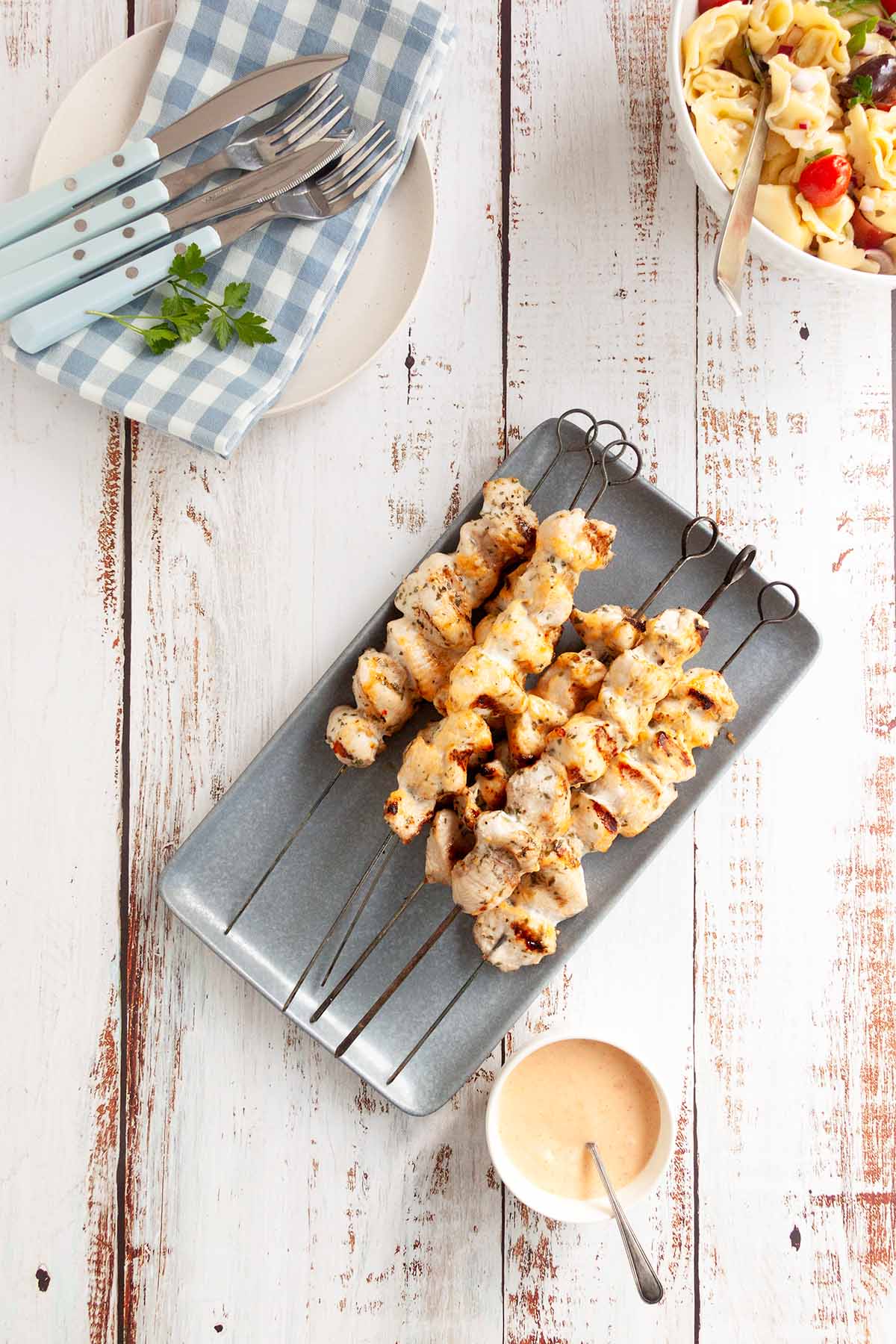 Grilled chicken skewers served on a modern slate tray with creamy sauce on the side and a fresh pasta salad in the background, ready for a delightful meal.