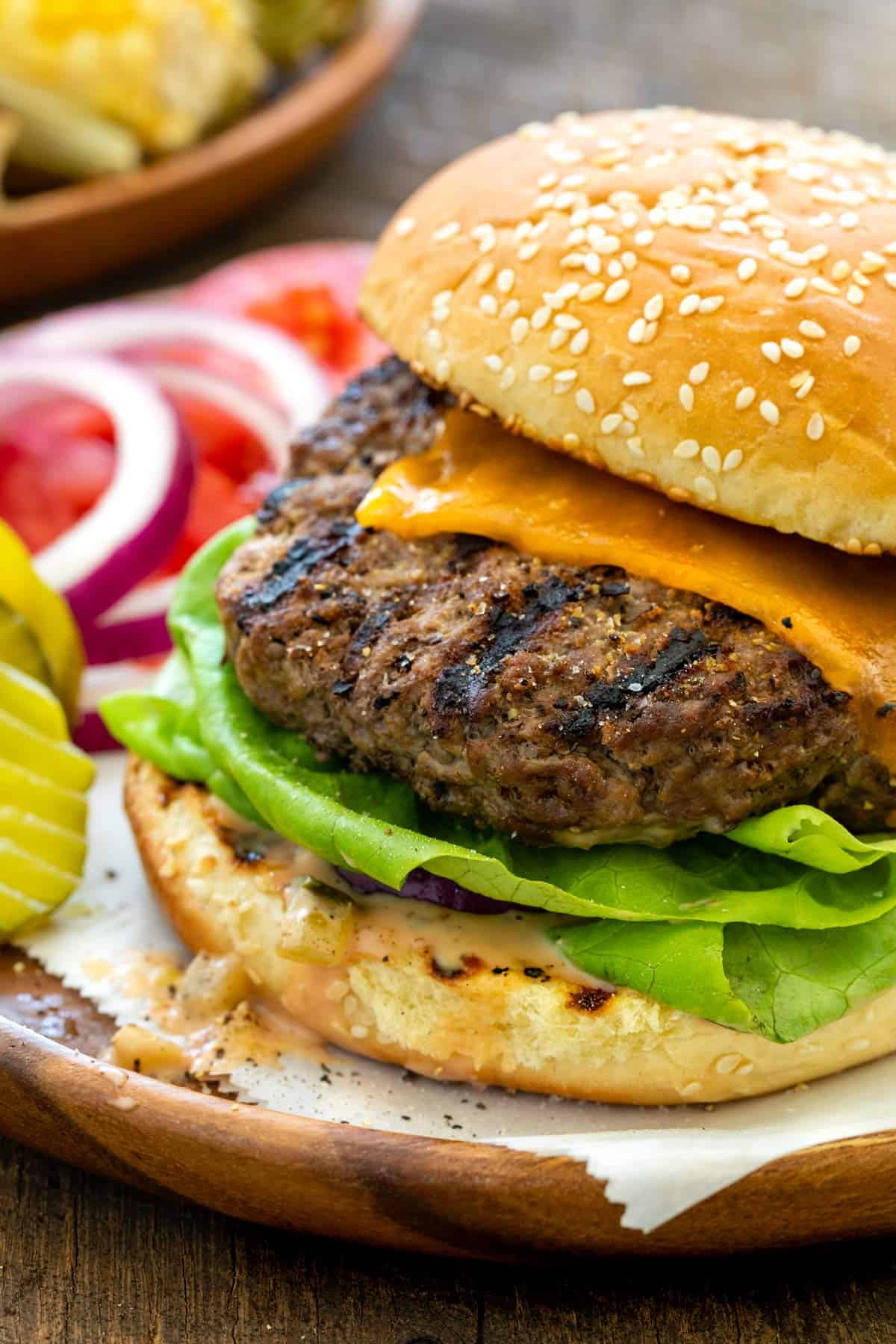 A grilled cheeseburger with lettuce and onions on a sesame seed bun, served with pickles and sliced onions on a wooden plate.