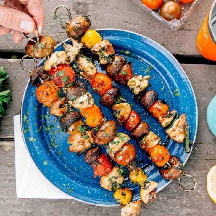 Four kabobs with chicken and vegetables on a blue plate. A hand is reaching in to pick one up.