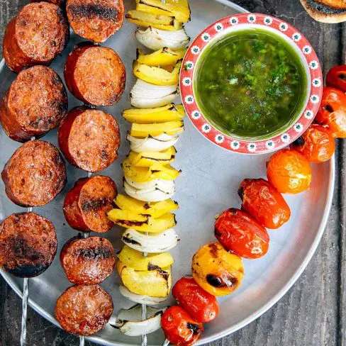 A vibrant bbq feast featuring juicy grilled sausage, colorful vegetable skewers with onions, peppers, and tomatoes, plus a tantalizing bowl of green sauce, all served on a rustic metal tray.