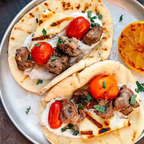Grilled skewers of succulent meat and roasted cherry tomatoes served on warm flatbreads with a dollop of creamy sauce, garnished with herbs and accompanied by a charred lemon half, suggesting a zesty flavor note.