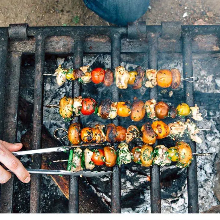 Grilling colorful skewers of mixed vegetables over an open flame, with a person using tongs to turn them for even cooking.