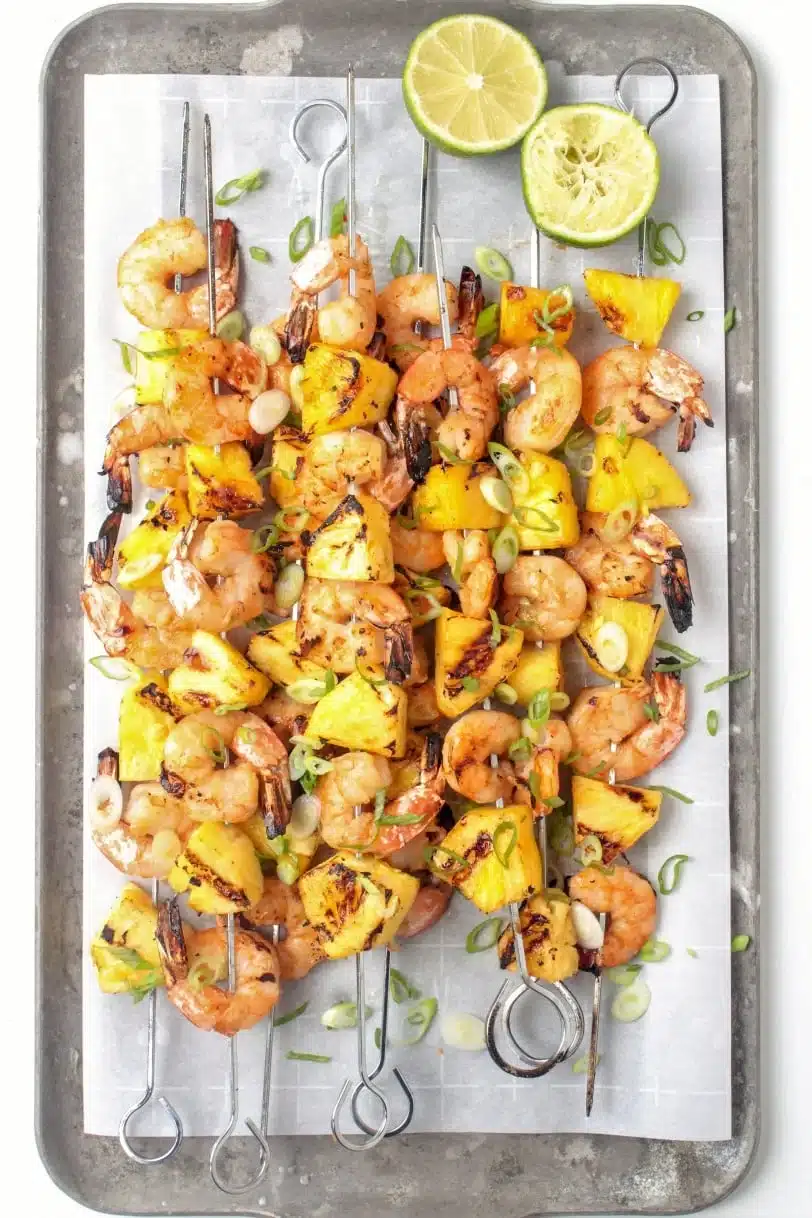 Grilled shrimp and pineapple skewers on a metal tray garnished with lime and sliced green onions.
