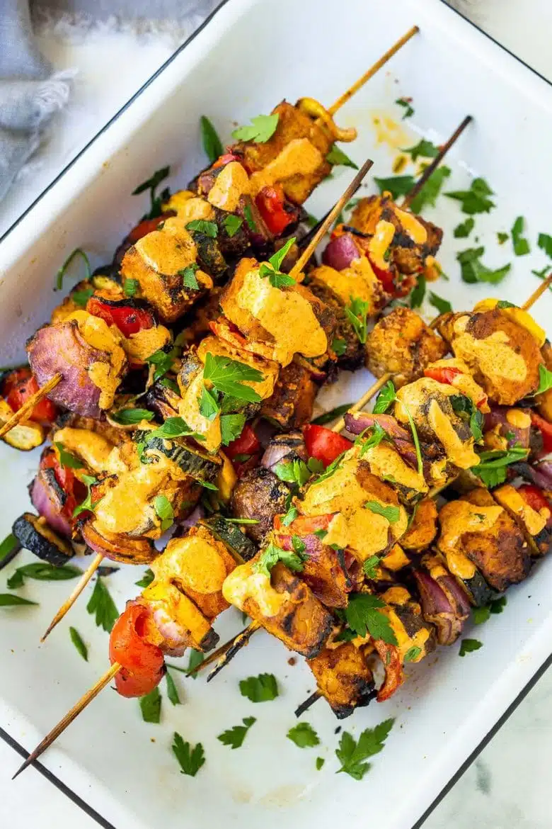 Colorful, grilled vegetable and tofu skewers garnished with fresh herbs and crispy chips, served on a white platter.
