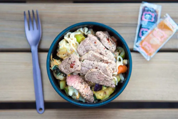 A bowl of noodle salad topped with grilled meat and vegetables, accompanied by a fork and condiment packets on a wooden table.