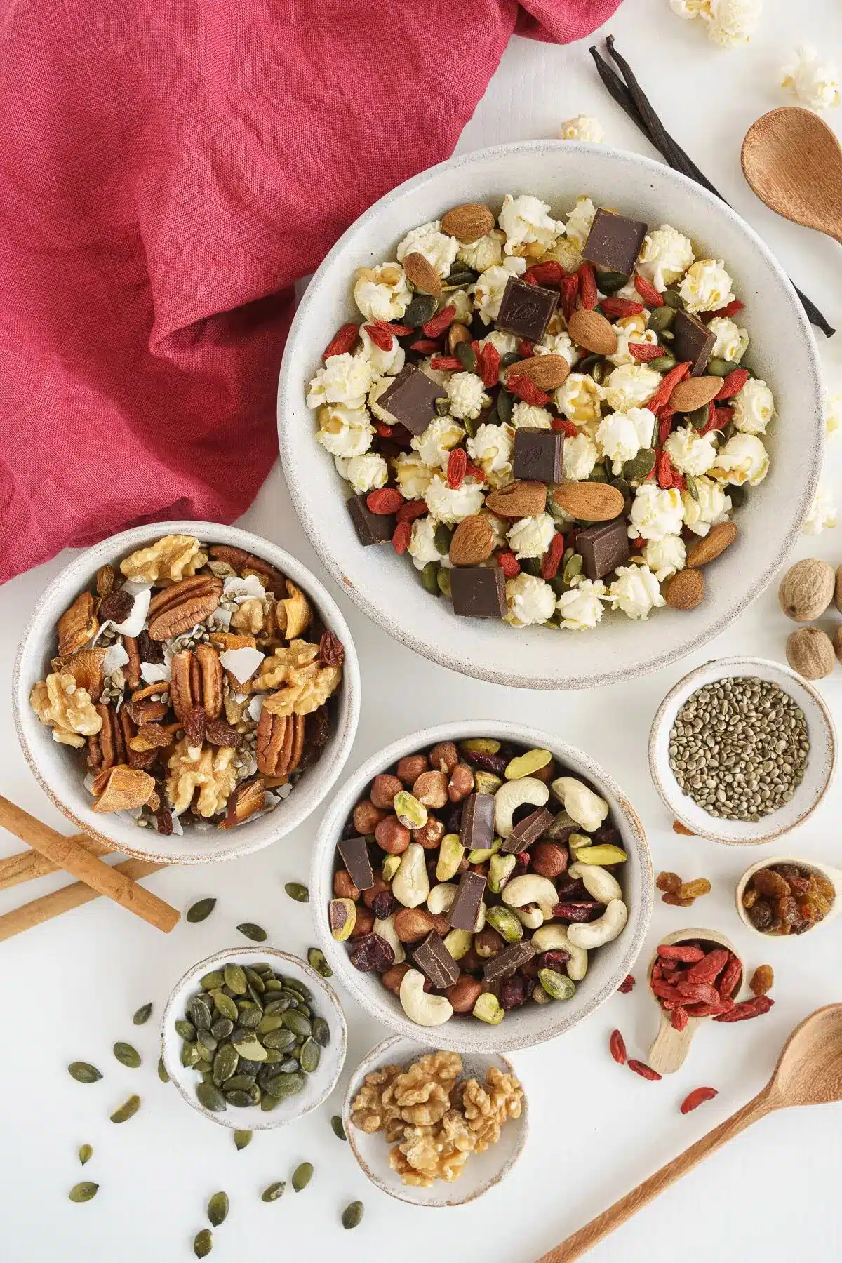 Bowls of trail mix, nuts, seeds, and popcorn are arranged on a white surface, accompanied by wooden spoons and a red cloth.