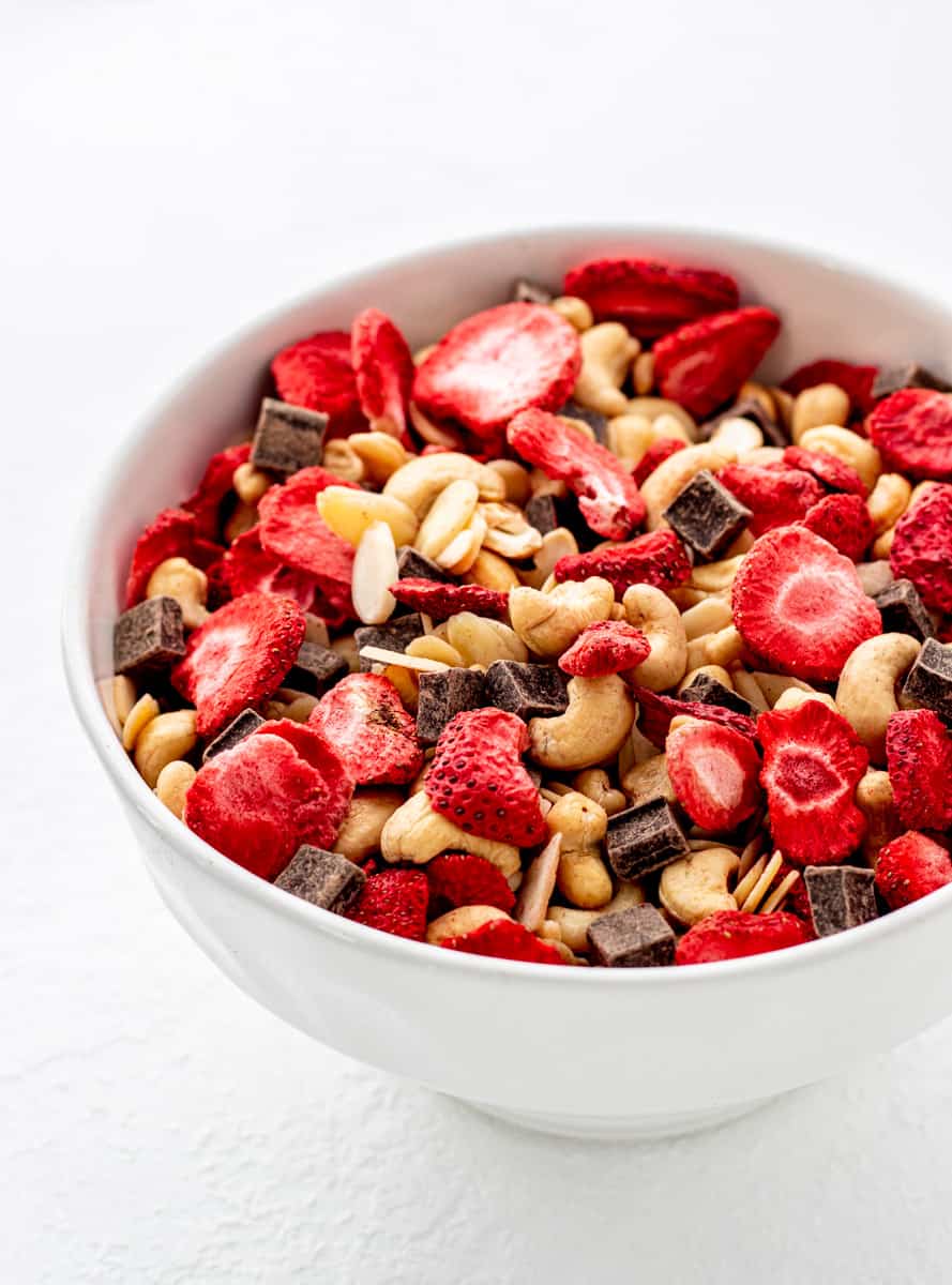 A white background hosts a bowl filled with trail mix, featuring dried strawberries, nuts, and chocolate pieces.
