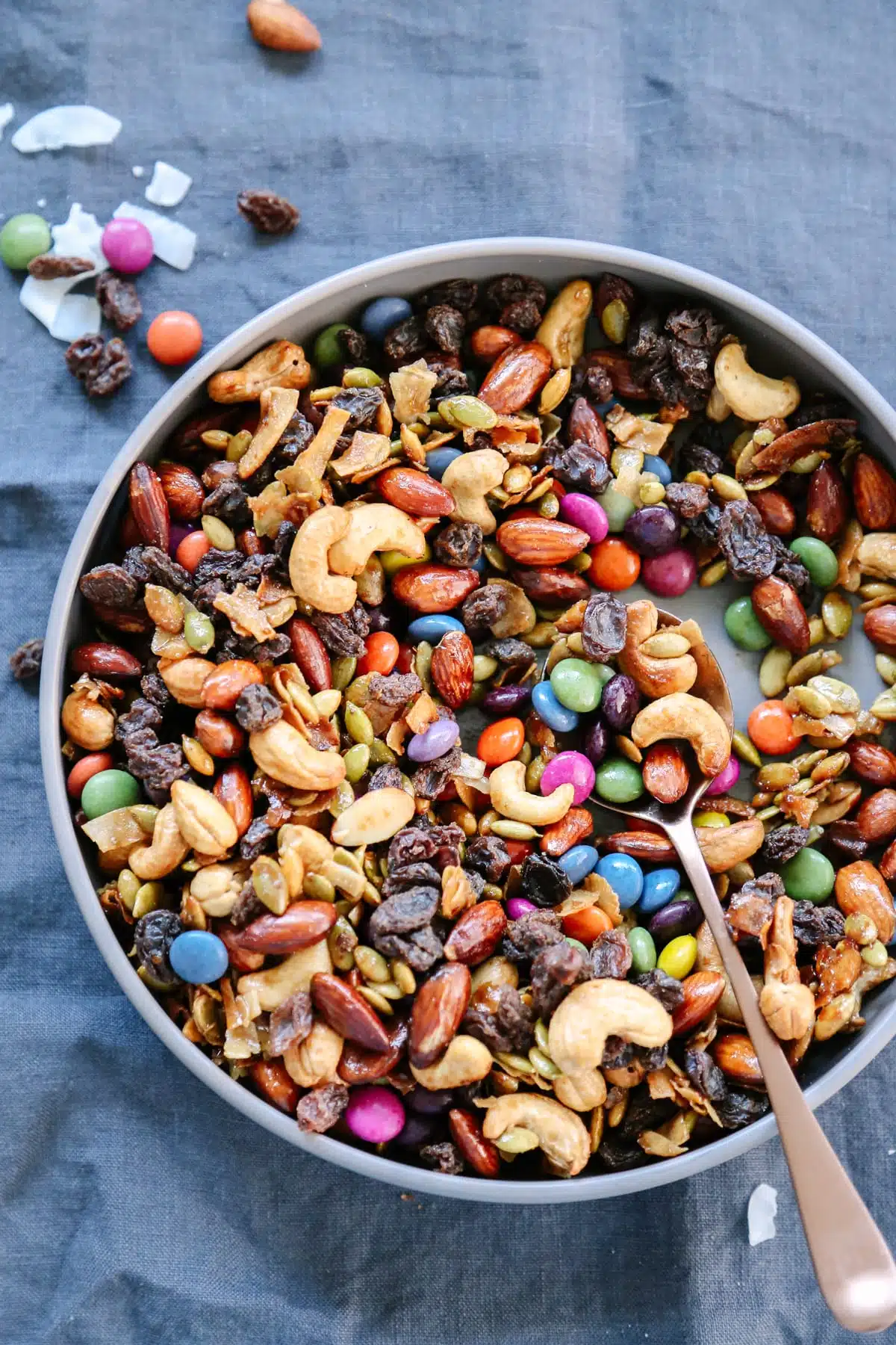 A bowl of trail mix with cashews, almonds, raisins, m&m's, pistachios, and walnuts is placed on a dark grey fabric surface.