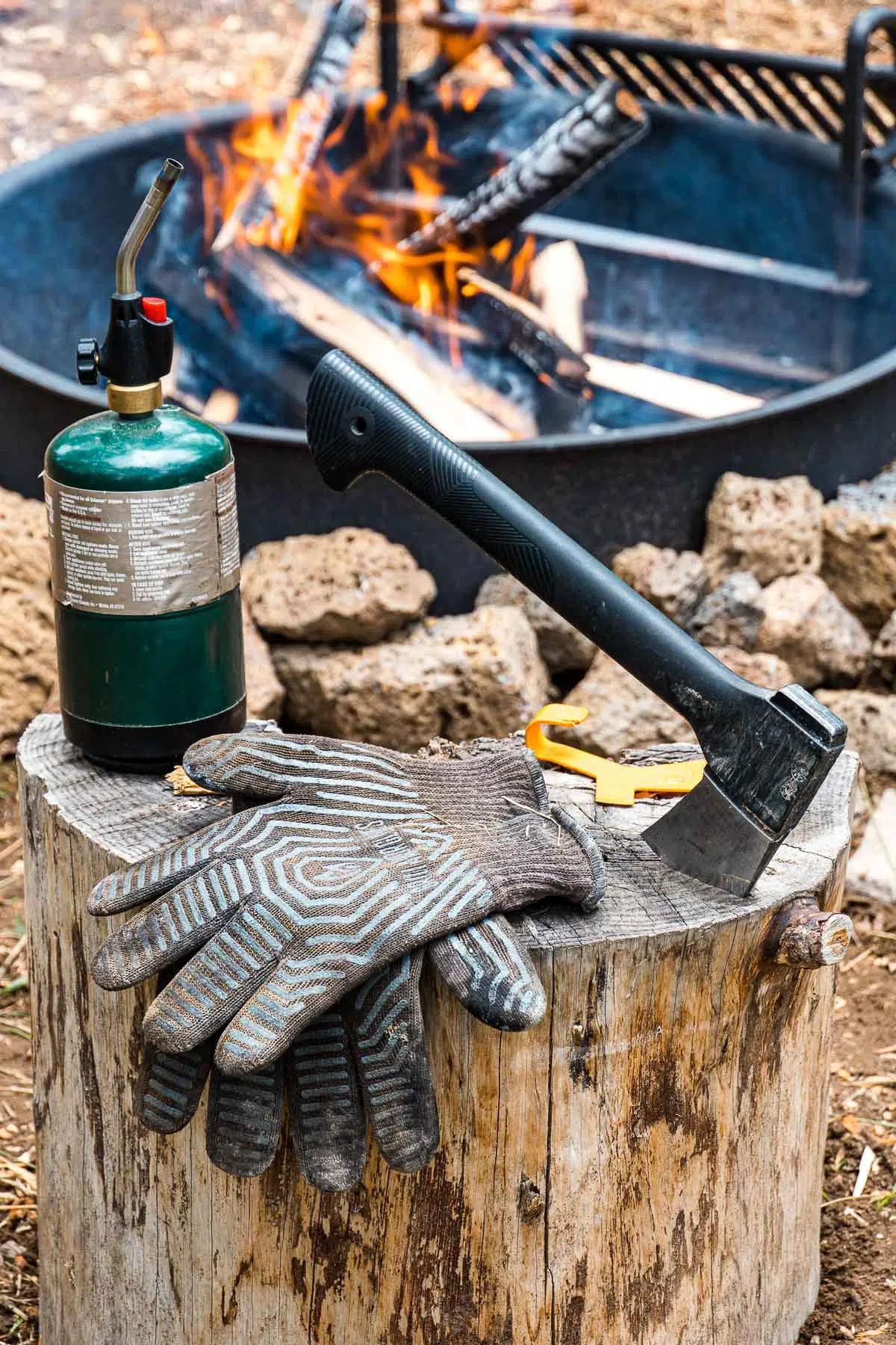Torch, hatchet, and heat proof gloves arranged on a log with a campfire in the background