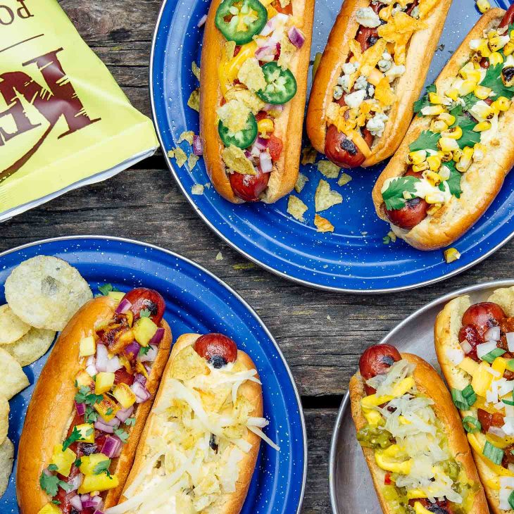 Three blue plates with hotdogs in buns and various toppings