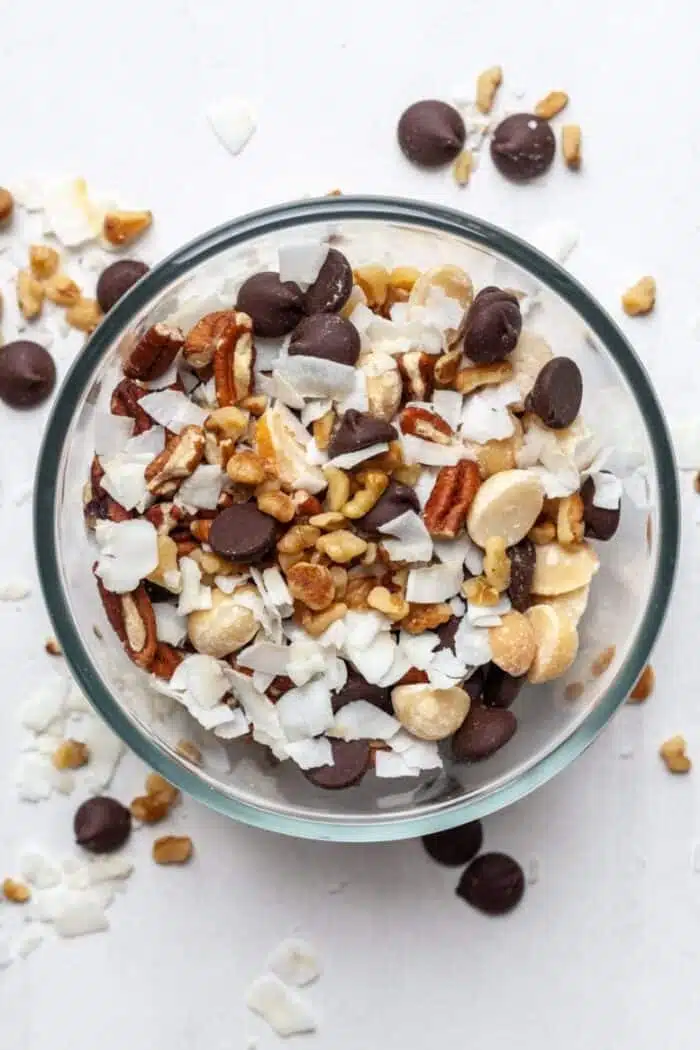 A bowl is filled with mixed trail mix including chocolate chips, pecans, coconut shavings, and peanuts against a white backdrop.