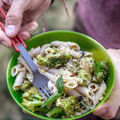 A person eating a healthy broccoli and pasta salad from a green bowl with a fork, outdoors.