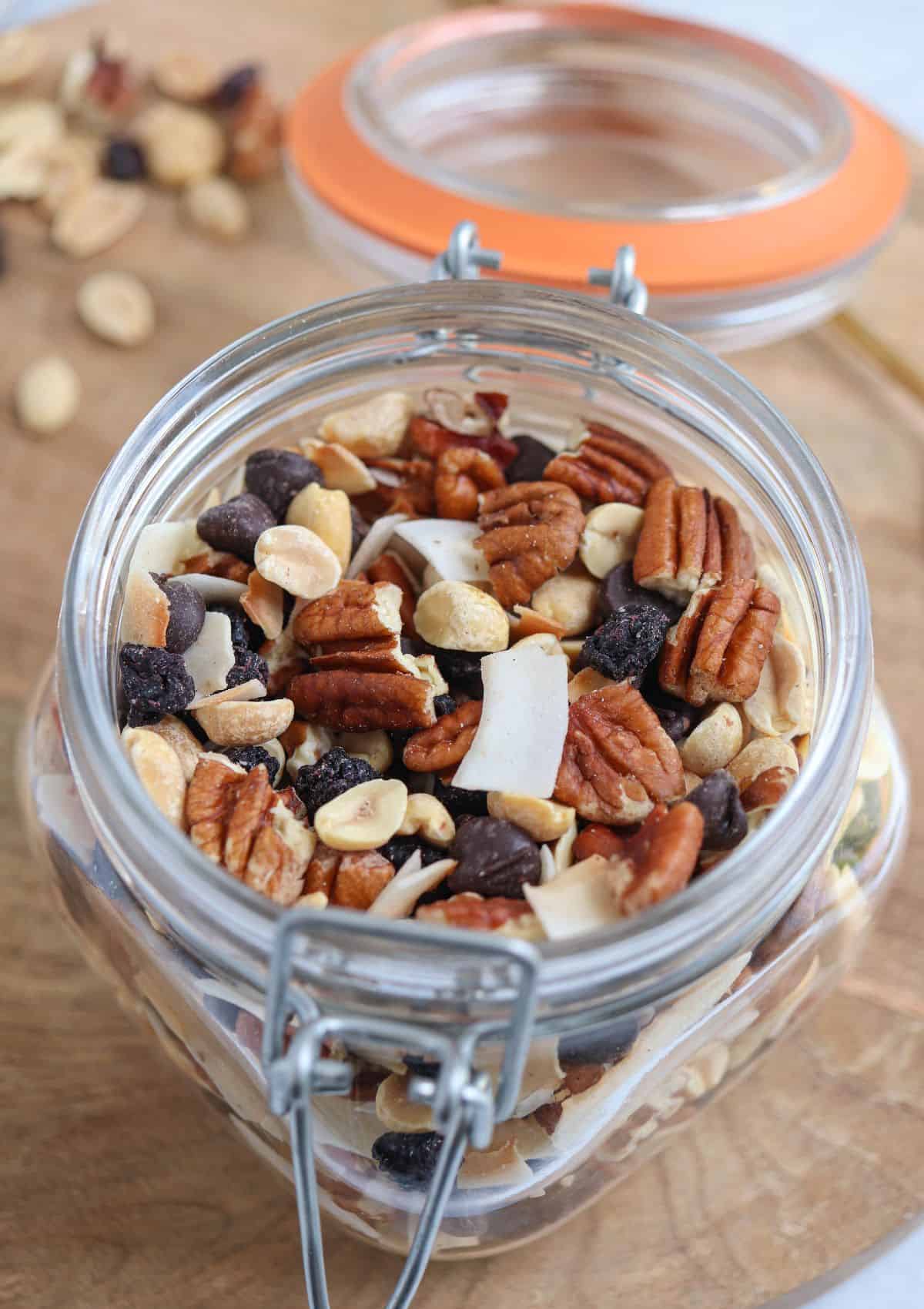 A glass jar on a wooden surface contains a blend of nuts, dried fruits, and chocolate chips, surrounded by scattered mix and an open orange lid.