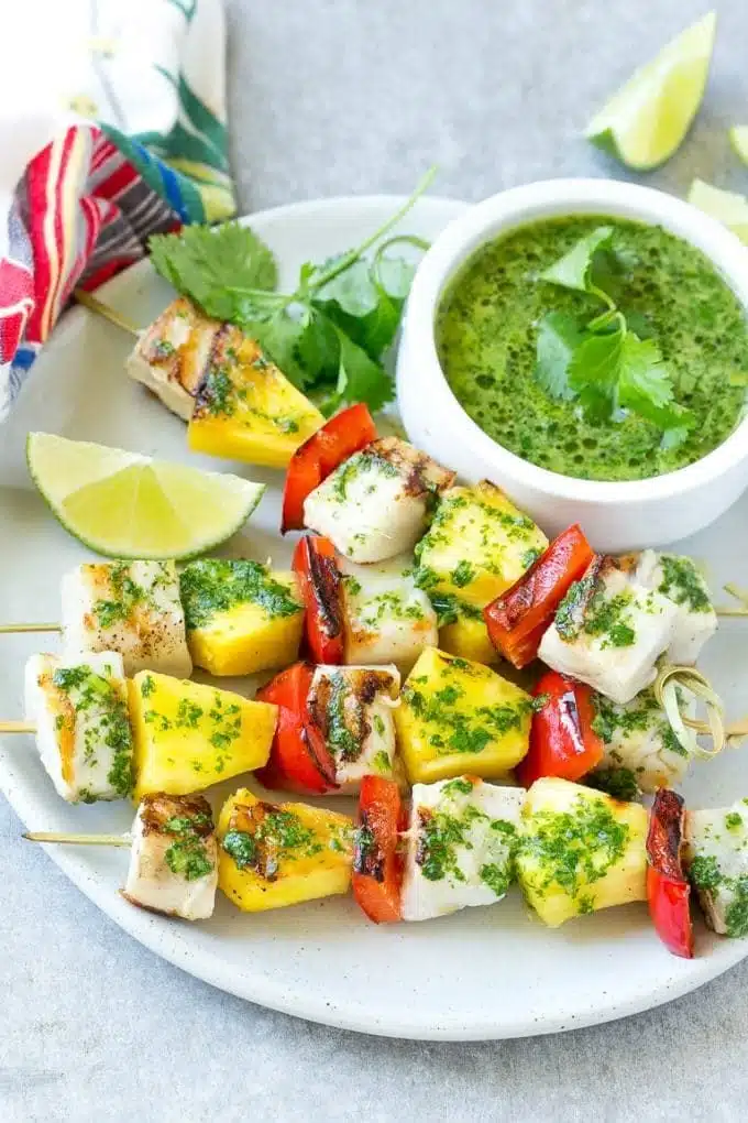 Grilled vegetable skewers with zucchini, red bell peppers, and onions, served with a side of fresh green herb sauce and lime wedges on a white plate, ready to be enjoyed as a healthy and colorful meal.