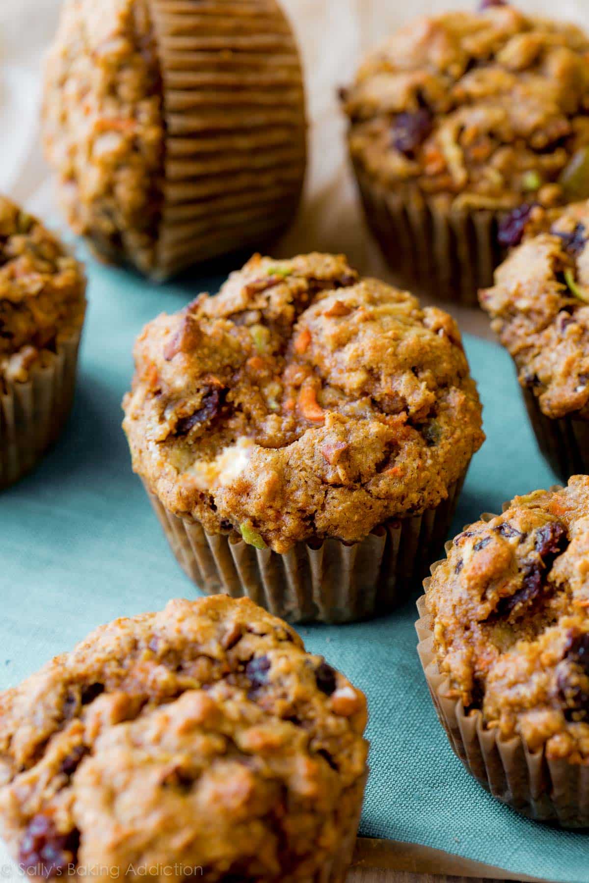 Freshly baked muffins with visible fruit and nut pieces are arranged on a teal napkin against a soft-focus background.