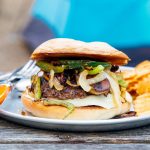 A burger topped with green peppers and onions on a blue plate and camping scene in the background