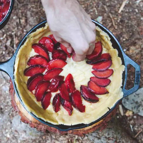 Arranging plum slices on a tart in a cast iron pan outdoors, with a hand in motion placing the fruit.