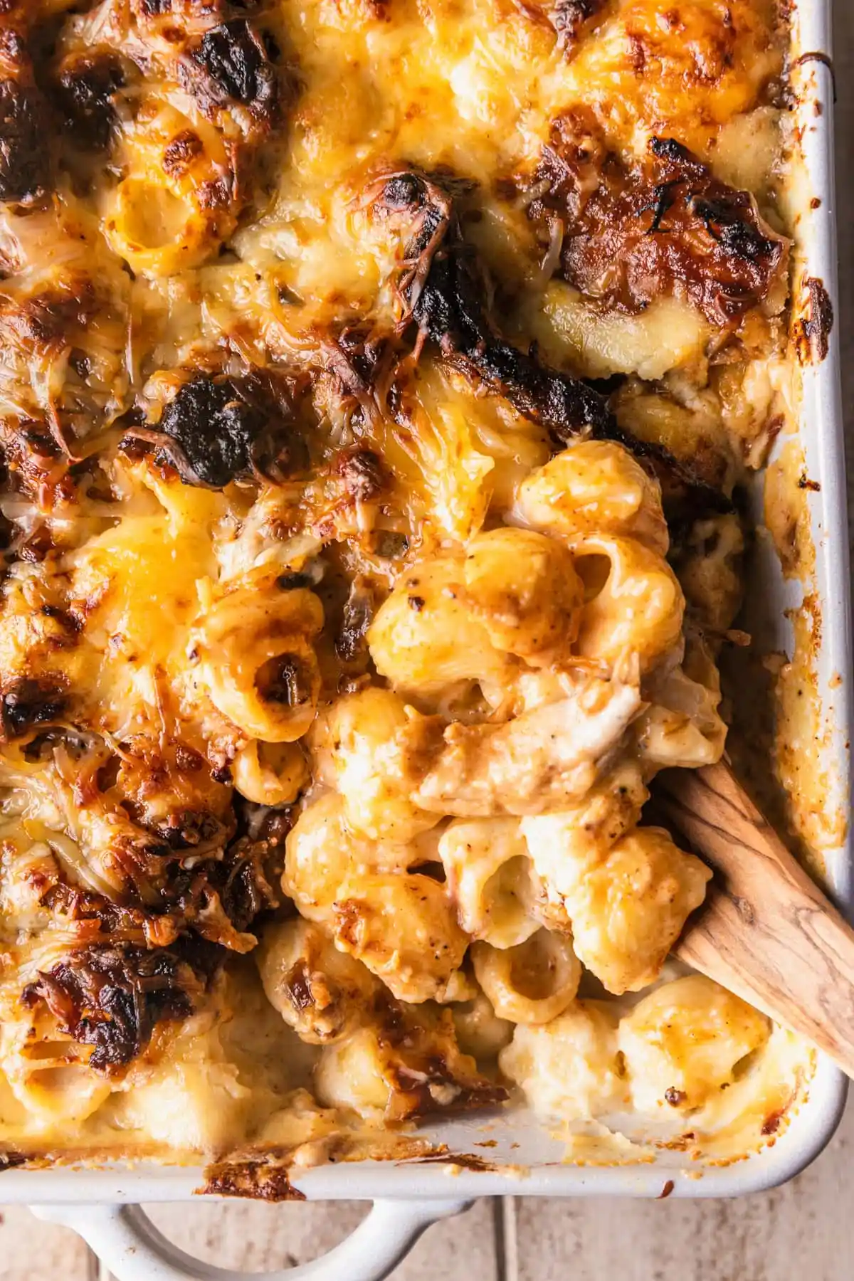 A baked macaroni and cheese dish in a white casserole has a golden-brown cheese crust, accompanied by a wooden spoon.