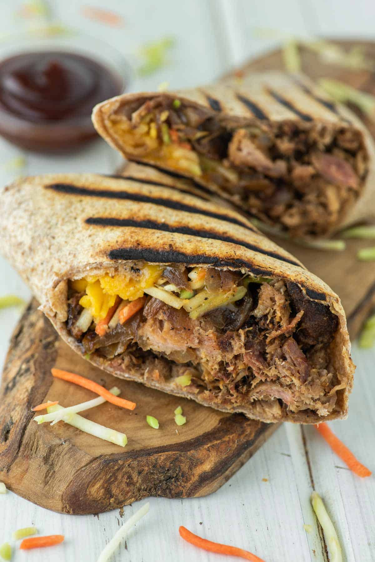 Two halved grilled wrap sandwiches containing shredded meat and vegetables are situated on a wooden board, accompanied by a side of sauce and sprinkled with finely chopped vegetables.