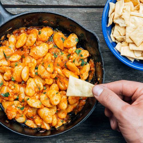 Hand holding a pita chip with Spanish White Bean Dip in a cast iron skillet