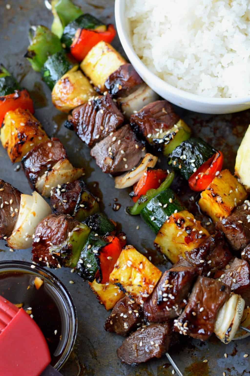 Grilled beef and vegetable skewers with pineapple chunks served next to a bowl of white rice, sprinkled with sesame seeds on a rustic metal surface.