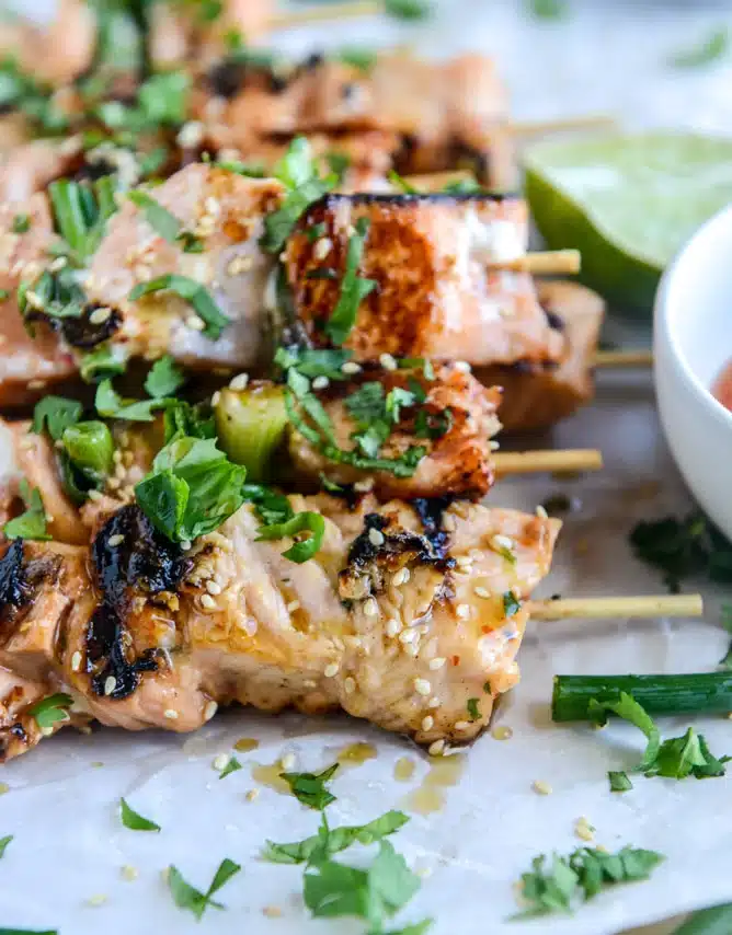 Grilled chicken skewers garnished with sesame seeds and chopped herbs, served with a lime wedge and dipping sauce on the side.