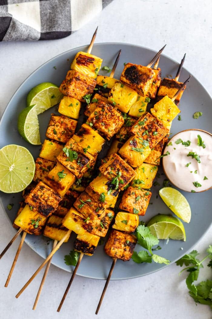 Grilled pineapple and tofu skewers are adorned with fresh cilantro and lime wedges, accompanied by a side of creamy dipping sauce.