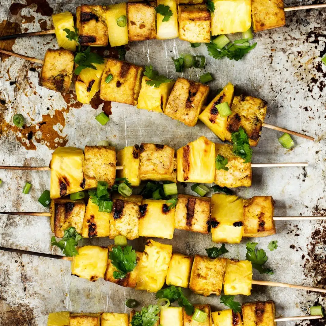 Grilled pineapple and tofu skewers seasoned with herbs on a rustic metal surface, showcasing a delicious vegan bbq option.