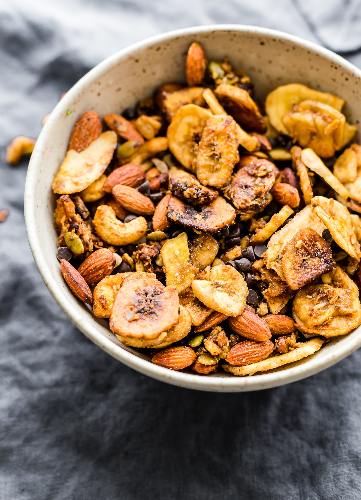 A bowl of homemade trail mix with banana chips, almonds, and chocolate chunks is close-up on a textured grey cloth.