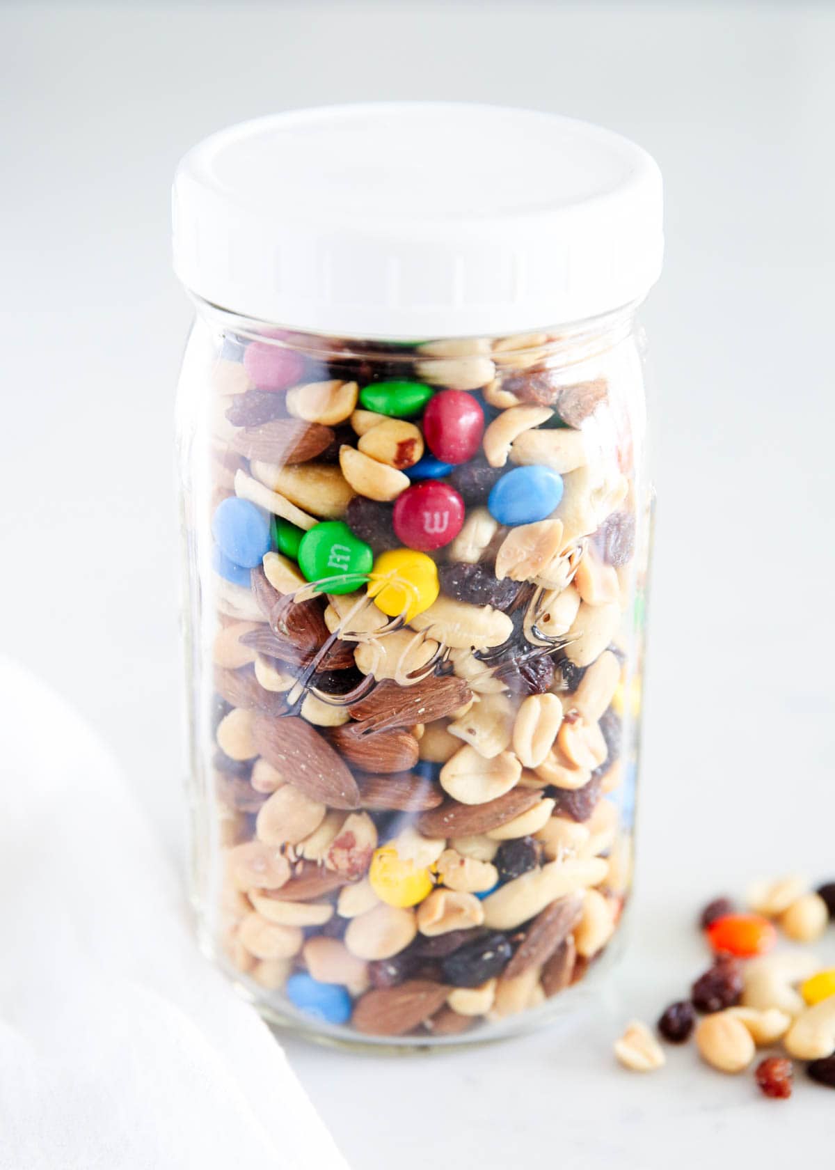 A clear glass jar with a white lid houses an assortment of mixed nuts, seeds, and colorful M&M candies on a light backdrop.