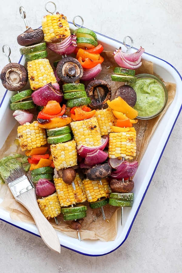 Colorful vegetable skewers with corn, zucchini, peppers, onions, and mushrooms, served with a side of green sauce in a white baking dish, ready for grilling or serving.