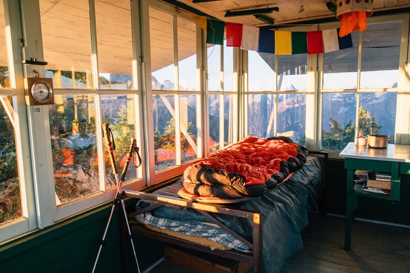 The interior of a fire lookout, with a sleeping bag on a cot and windows looking out to a view of mountain peaks.